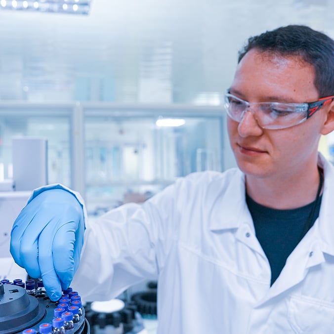 Un technicien de laboratoire portant une blouse blanche, des lunettes de protection et des gants bleus touche de petites fioles alignés en rond.