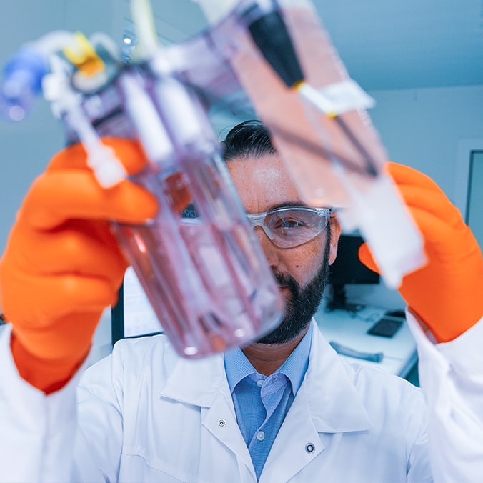 A laboratory technician wearing a white coat and protective goggles lifts a utensil with his orange gloves.