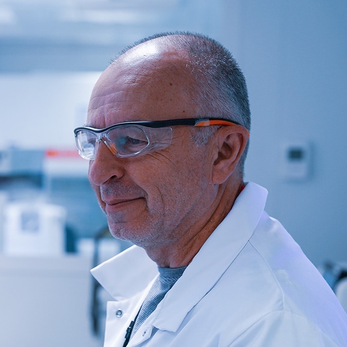 A lab technician wearing goggles and a white coat smiles.