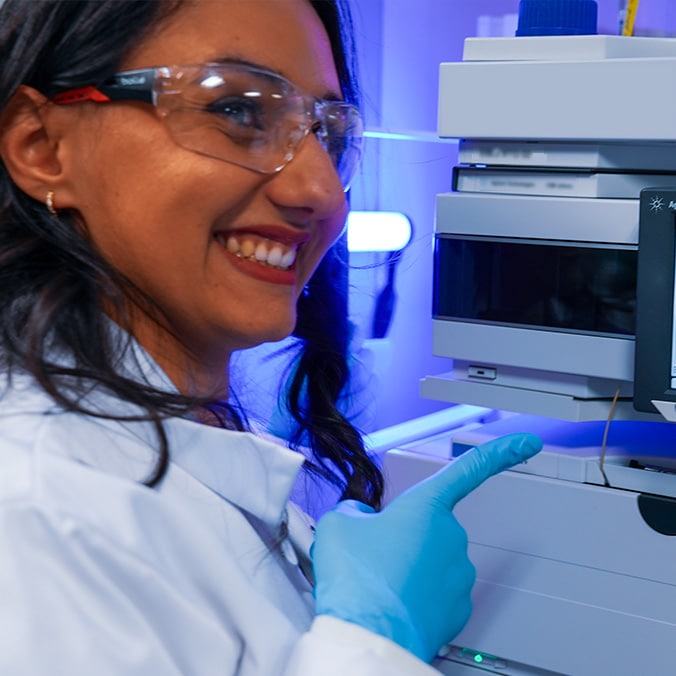 A lab technician wearing goggles, a white lab coat and blue gloves smiles as she points at a machine with her finger.