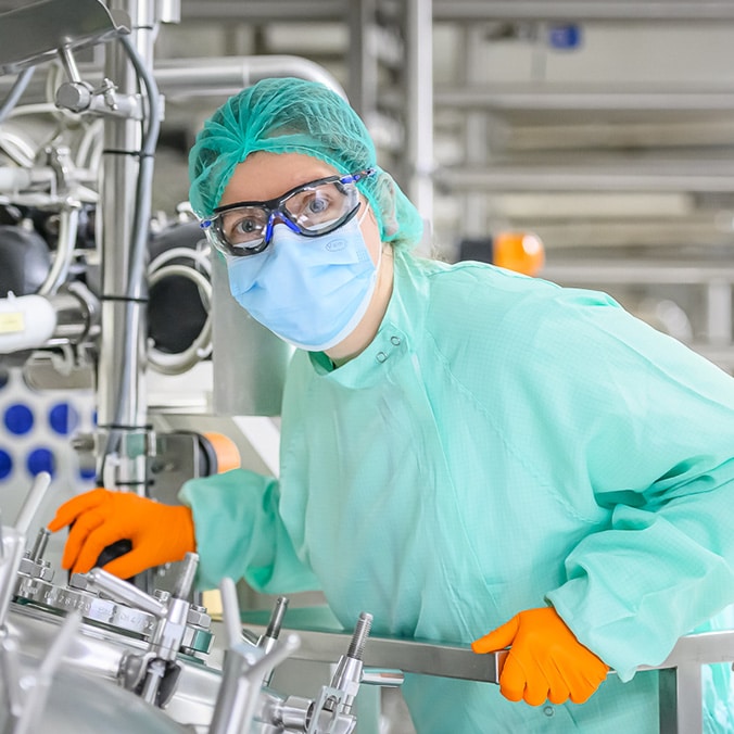 A laboratory technician wears a green lab coat, surgical mask, goggles and a hair cap. He operates a machine wearing orange gloves for added safety.