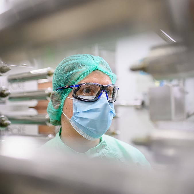 Un technicien dans un laboratoire portant une charlotte verte, des lunettes de protection, un masque chirurgical.