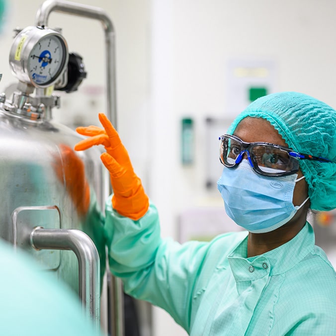 A lab technician dressed in a green lab coat, green cap and goggles places his orange-gloved hand on a machine.