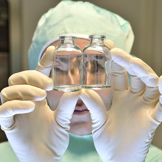 A lab technician wearing a green lab coat, white cap and gloves lifts two small clear glass bottles in front of his eyes.