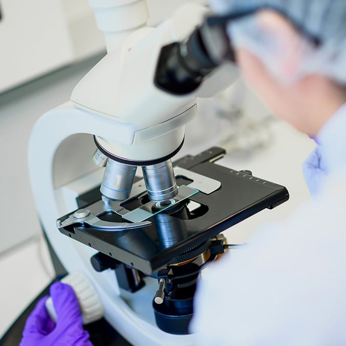 Un technicien de laboratoire portant une charlotte et des gants analyse une substance indéfinie au microscope.